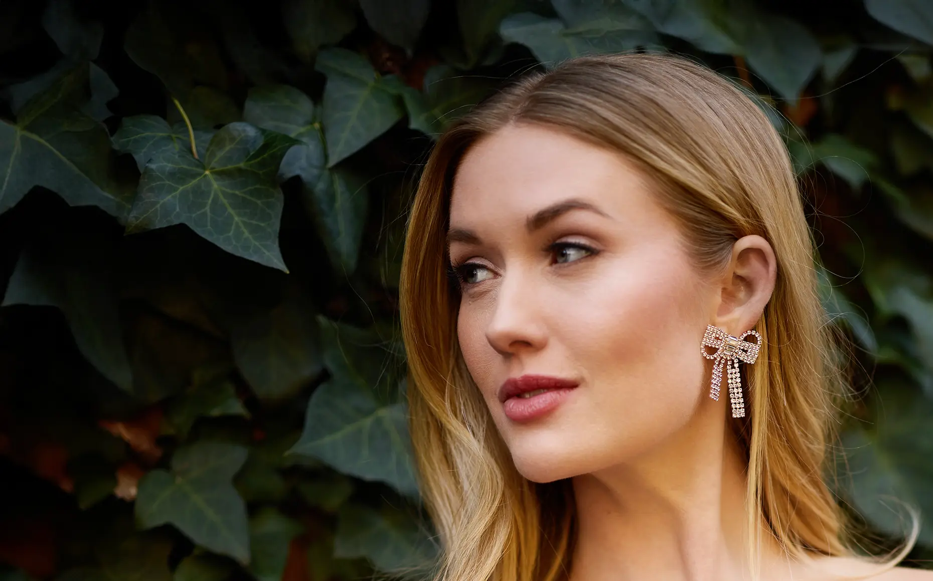 Model wearing a crystal bow drop earring against a backdrop of ivy leaves